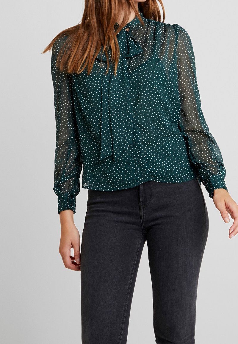 Blusa verde a pois con collo alto, maniche lunghe trasparenti e dettaglio a fiocco al colletto, abbinata a jeans scuri.