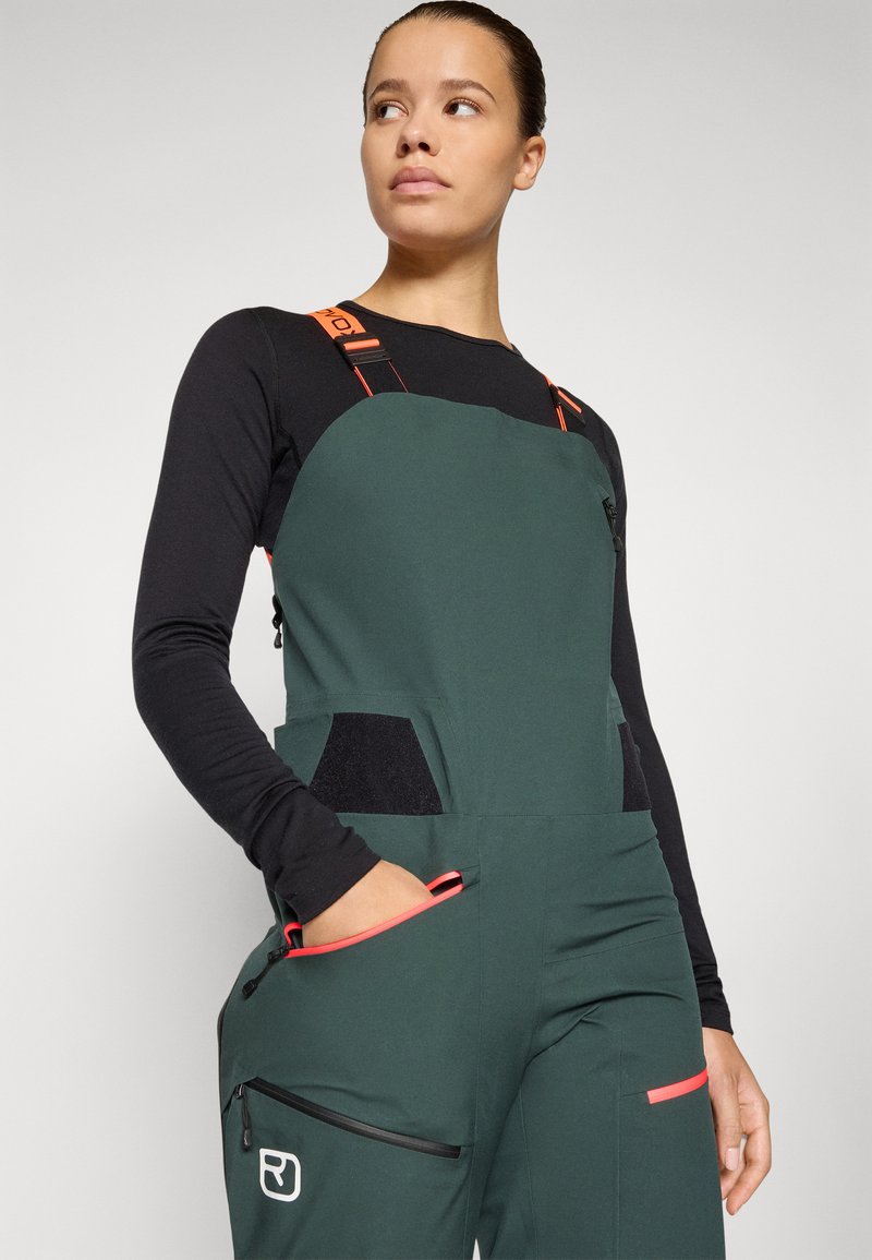 Grüne Latzhose mit schwarzen Akzenten, ausgestattet mit verstellbaren orangefarbenen Trägern, Reißverschlusstaschen und einer glatten Textur. Elegantes, modernes Design.