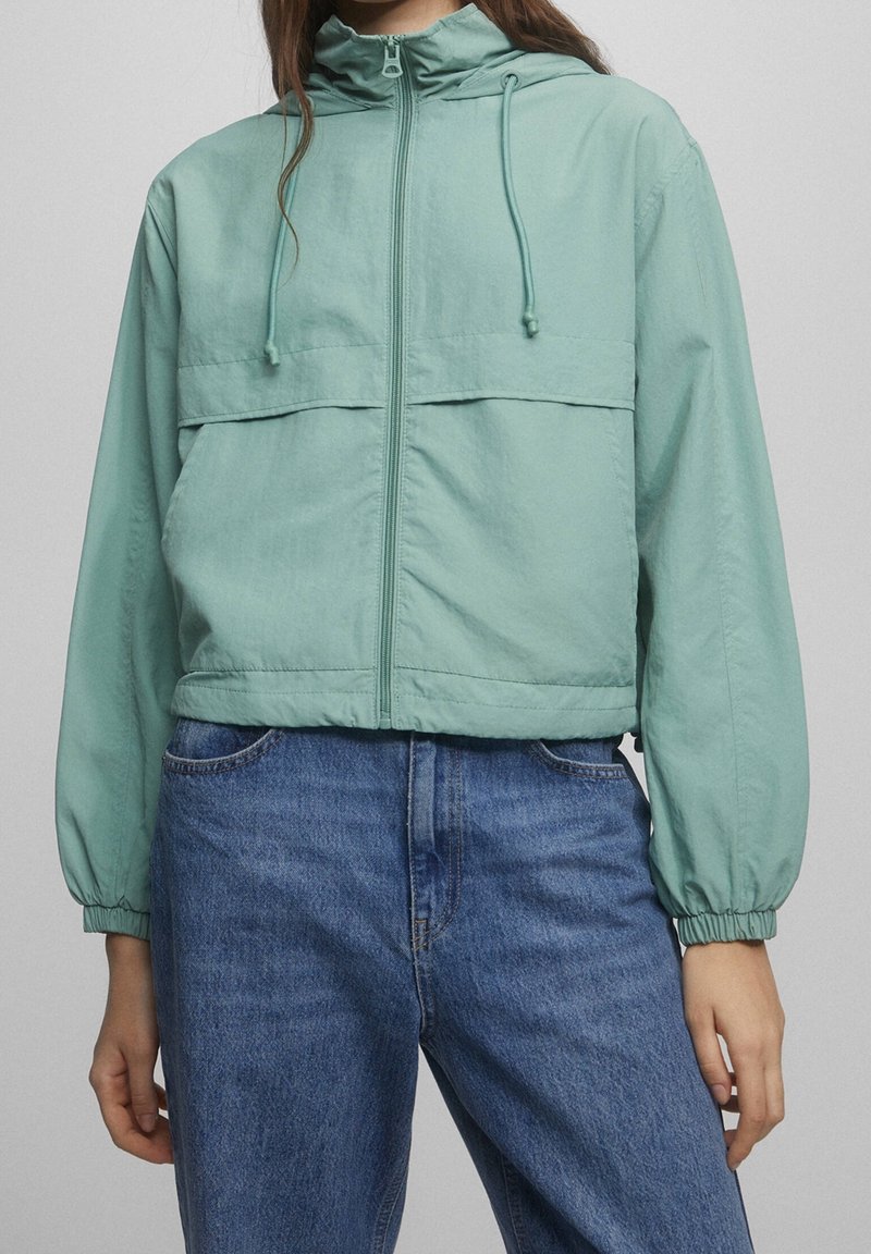 Kvinna i ljusgrön huvtröja med dragkedja och elastiska manschetter, samt blå denimjeans, står mot en enfärgad bakgrund.