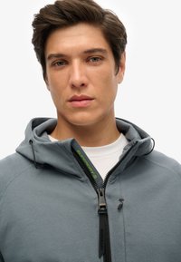 Sudadera ligera gris con capucha, que presenta una cremallera negra y un logo verde. Textura suave con un diseño ajustado y costuras en contraste.