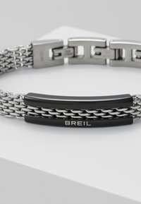 Ruostumatonta terästä oleva rannekoru, jossa on kudottuja ketjuliitoksia ja musta korostustanko, johon on kaiverrettu "BREIL". Suorapintainen rakenne ja hopean värinen viimeistely.