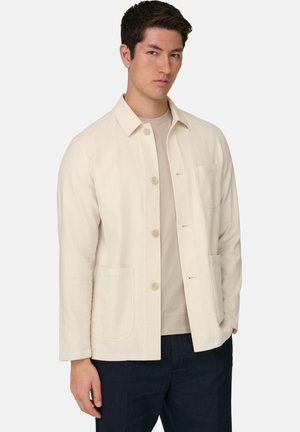 Uomo che indossa una giacca color beige con bottoni e colletto sopra una camicia beige chiaro e pantaloni scuri, in piedi davanti a uno sfondo neutro.