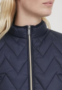 Giacca trapuntata blu navy con colletto alto, dotata di una cerniera argento e un motivo a zig zag sul tessuto, texture liscia.