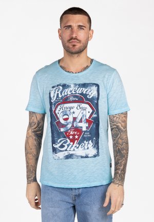 Mann mit Tätowierungen, der ein hellblaues T-Shirt mit der Grafik "Raceway Bikers 94" und blaue Jeans trägt, steht vor weißem Hintergrund.