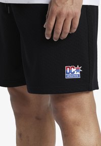 Pantaloni sportivi neri realizzati in tessuto a rete, con un logo ricamato "DC Athletics" in rosso, bianco e blu sulla coscia sinistra.