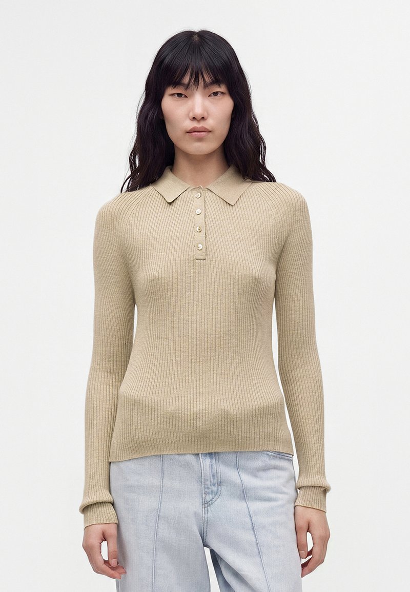 Femme aux longs cheveux foncés portant un pull polo côtelé beige à manches longues et un jean taille haute bleu clair, debout devant un fond uni.
