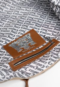 Etichetta in pelle marrone con logo argento in rilievo su una fodera bianca, caratterizzata da motivi in testo nero. Include una tasca con zip e cerniera nera.