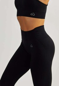 Schwarzes Sportbekleidungsset mit einem kurzen Oberteil, das ein kleines Herz-Logo zeigt, und hochgeschnittenen Leggings aus strukturiertem Material mit einem ähnlichen Logo.
