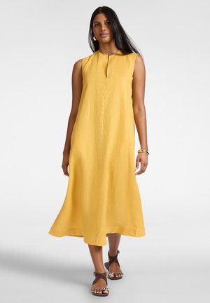 Kobieta w żółtej sukience bez rękawów o długości do połowy łydki, z dekoltem typu keyhole, zestawiona z brązowymi sandałami na paski i złotą biżuterią.