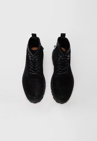 Botas de tobillo de ante negro con cierre de cordones en la parte delantera, detalles de cremallera y suela de goma. Presentan una textura suave y costuras reforzadas.