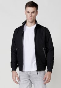 Chaqueta negra con cremallera, cuello alto, bolsillos frontales con cremallera, puños elásticos y sutil detalle del logo en el pecho. Llevada sobre una camiseta blanca.