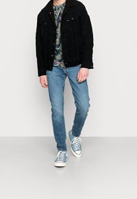 Man som bär en svart kordjacka, blommig skjorta, ljusblå jeans och ljusblå canvas-sneakers som går mot en enkel bakgrund.