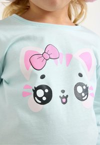 Vaaleansininen puuvillainen t-paita, jossa on painettu kissan kasvot suurine silmineen, vaaleanpunainen rusetti ja viiksikoristeet. Yksinkertainen, leikkisä muotoilu.