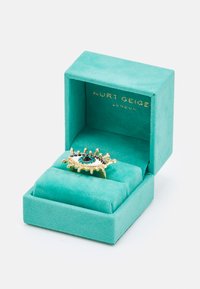 Złotą pierścionek z wzorem oka, ozdobiony niebieskimi, czarnymi i przezroczystymi kamieniami, umieszczony w jasnoniebieskim pudełku z zamszowym wykończeniem.