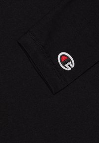 Maglietta nera a maniche lunghe in morbido tessuto. Presenta un piccolo logo ricamato con un accento rosso sul polsino. Design semplice e senza decorazioni.