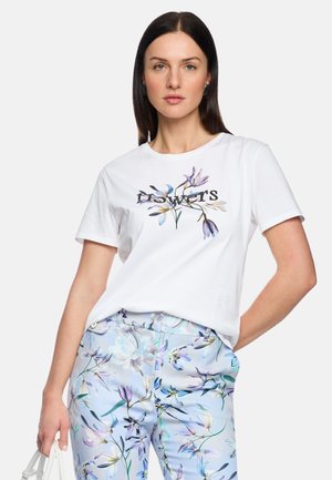 Frau trägt weißes T-Shirt mit floralem "Flowers"-Aufdruck und hellblaue Blumenhose, hält eine weiße Handtasche, vor einfachem Hintergrund.