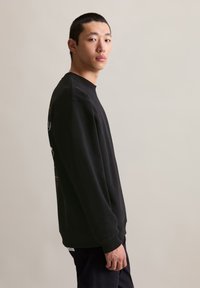 Felpa nera con collo rotondo e maniche lunghe, caratterizzata da dettagli grafici sui fianchi. Indossata con pantaloni scuri, mostrata di profilo.
