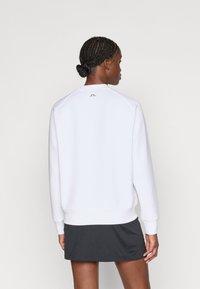 Biely sveter s dlhými rukávmi, s okrúhlym výstrihom a zlatým logom na chrbte, skombinovaný s tmavošedou sukňou. Hladká textúra.