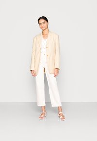 Blazer bege, top branco com rendas e calças brancas cortadas. Modelo está em pé sobre um chão cinza, usando sandálias brancas de tiras. Design simples, cores claras.