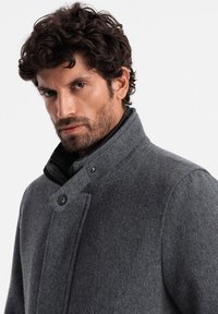 Abrigo de lana gris con un alto cuello, que presenta un cierre de botones, un tejido texturizado y un diseño sofisticado.