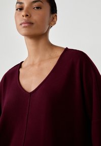 Burgundowy sweter z dekoltem V, wykonany z miękkiego materiału, o luźnym kroju, teksturowanym wykończeniu i widocznych detalach szwów.