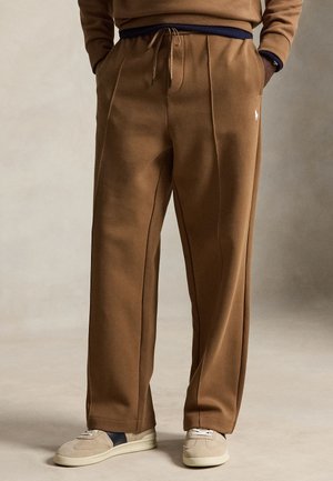 Pantalon de survêtement marron sur mesure avec taille à cordon et logo brodé, porté avec des baskets beige et bleu marine sur un fond neutre.