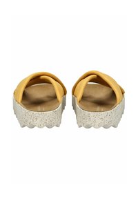 Žluté látkové slip-on boty se širokými křížovými pásky, položené na tečkované bílé platformě s vykrojenými okraji.