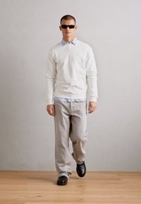 White sweater over a light blue collared shirt, gray pants, and black shoes. The model wears dark sunglasses and stands against a neutral background.