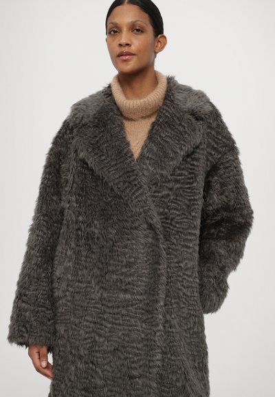 Pilkas dirbtinio kailio paltas su tekstūru, plačiu išpjautu apykaklė ir laisvu siluetu. Užsidėtas virš šviesiai rudos spalvos megztinio su aukštu kaklu.