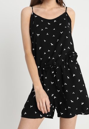 Nainen pukeutuneena mustaan hihattomaan haalariin, jossa valkoisia lintukuvioita ja kiristysnyöri vyötäröllä, seisomassa vaaleaa yksiväristä taustaa vasten.