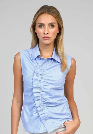 Femme aux cheveux blonds lisses portant un chemisier sans manches bleu clair à rayures avec des boutons froncés sur le devant, posant avec une main dans la poche.