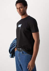 Camisa de algodón negra con un estampado gráfico, combinada con jeans de mezclilla azul y una chaqueta de mezclilla que cuelga del hombro.