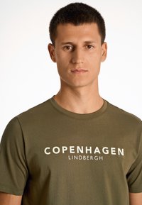 Camiseta de algodón verde oliva con "COPENHAGUE LINDBERGH" impreso en letras gruesas de color crema en el pecho; manga corta y escote redondo.