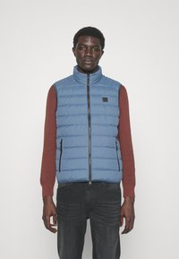 Světle modrá péřová vesta s vysokým límcem, horizontálním prošíváním a zipovými kapsami, nošená přes vínový svetr a tmavé džíny.