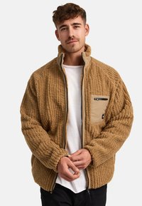 Chaqueta de forro polar texturizado marrón con cremallera frontal y cuello alto. Presenta un bolsillo con cremallera de color beis contrastante en el lado izquierdo del pecho.