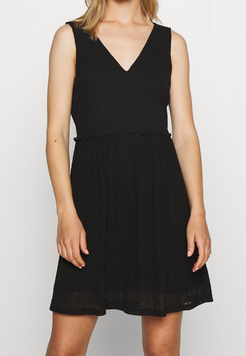Femme portant une robe noire sans manches à col en V, taille froncée et tissu texturé, debout devant un fond blanc uni.