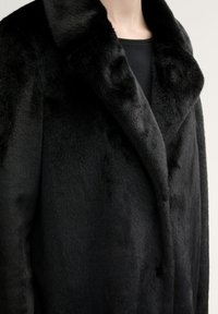 Cappotto in pelo finto nero con una consistenza morbida, colletto ampio e design a lunghezza completa. Caratterizzato da una silhouette classica e dettagli minimi.