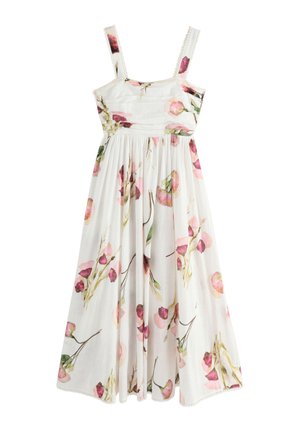 Jeune femme portant une robe blanche fleurie sans manches et des sandales, debout à l'extérieur sur une terrasse en pierre avec de l'herbe et un portail blanc derrière.
