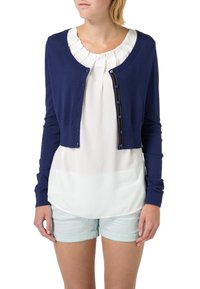 Námořnická modrá zkrácená kardigan s dlouhými rukávy, zapínáním na knoflíky a dekorativními detaily. Nosený přes krémovou blůzu a světlé šortky.