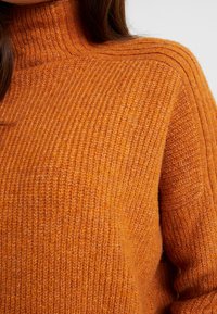 Dziany sweter w żywym pomarańczowym kolorze, z wysokim kołnierzem, fakturą prążkowaną i poziomymi paskami na ramionach.