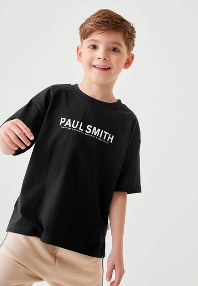 Melnas kokvilnas T-krekls ar īsām piedurknēm un apaļu kakla izgriezumu, uz kura priekšpuses ir balts "Paul Smith" logo. Gaiši bēšas bikses ar sānu svītrām.