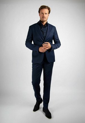 Man staat zelfverzekerd in een getailleerd marineblauw pak, zwart overhemd en zwarte schoenen, terwijl hij de knoop van zijn jasje rechtzet tegen een effen achtergrond.