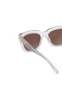 Kirkastettu kehysaurinkolasit ruskeilla linsseillä, kulmikas muotoilu ja läpinäkyvät templet. Kehyksessä on hienovarainen brändäys.