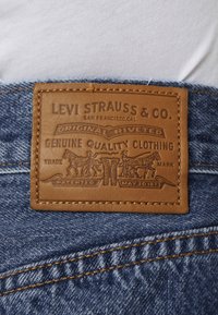 Étiquette en cuir brun cousue sur un denim bleu, présentant un texte en relief et un graphique de deux chevaux, des rivets et des dates de brevet.