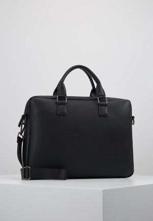 Mallette en cuir noir avec double poignées et bandoulière réglable, exposée sur un piédestal blanc sur un fond gris uni.