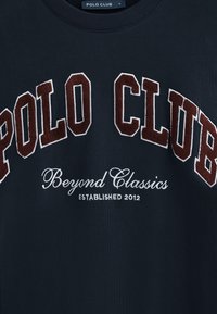 Marinblå sweatshirt med "POLO CLUB" i bordeaux och vit chenillebroderi, med "Beyond Classics" och "ETABLERT 2012" nedanför.