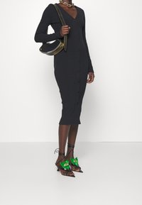 Femme portant une robe midi noire ajustée à boutons, des sandales à talons noires et vertes, un collier en or massif et tenant un sac à main noir.