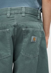 Πράσινο τζιν παντελόνι με πίσω τσέπες, η μία με έμπλαστρο λογότυπο Carhartt, φορεμένο από ένα άτομο με μπλε πουκάμισο.