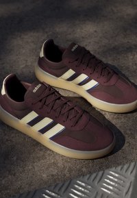 Paire de baskets Adidas bordeaux foncé avec des rayures blanches et bleues et des semelles couleur crème sur une surface en béton texturé.