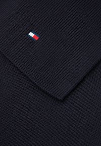 Marineblå strikket genser med ribbet tekstur; har en liten brodert logo i rødt, hvitt og blått.